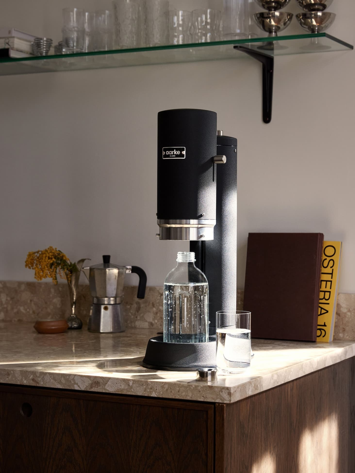 Aarke Carbonator Pro Matte Black on kitchen counter with a bottle of sparkling water, next to a crystal glass and coffee maker.