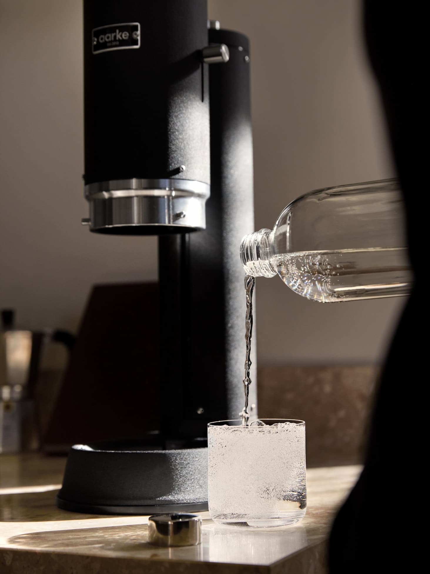 Sparkling water being poured from an Aarke bottle into a glass, next to an Aarke Carbonator Pro Matte Black on a kitchen counter.