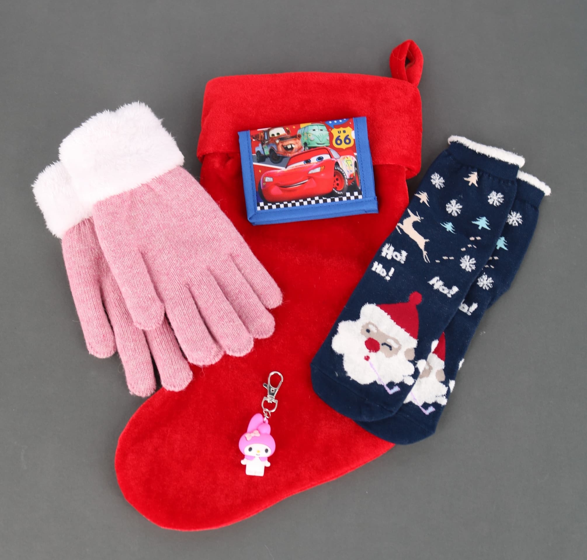A stocking with multiple items on top of it, including gloves, a child's Cars wallet, Santa Claus socks, and a Hello Kitty bag charm