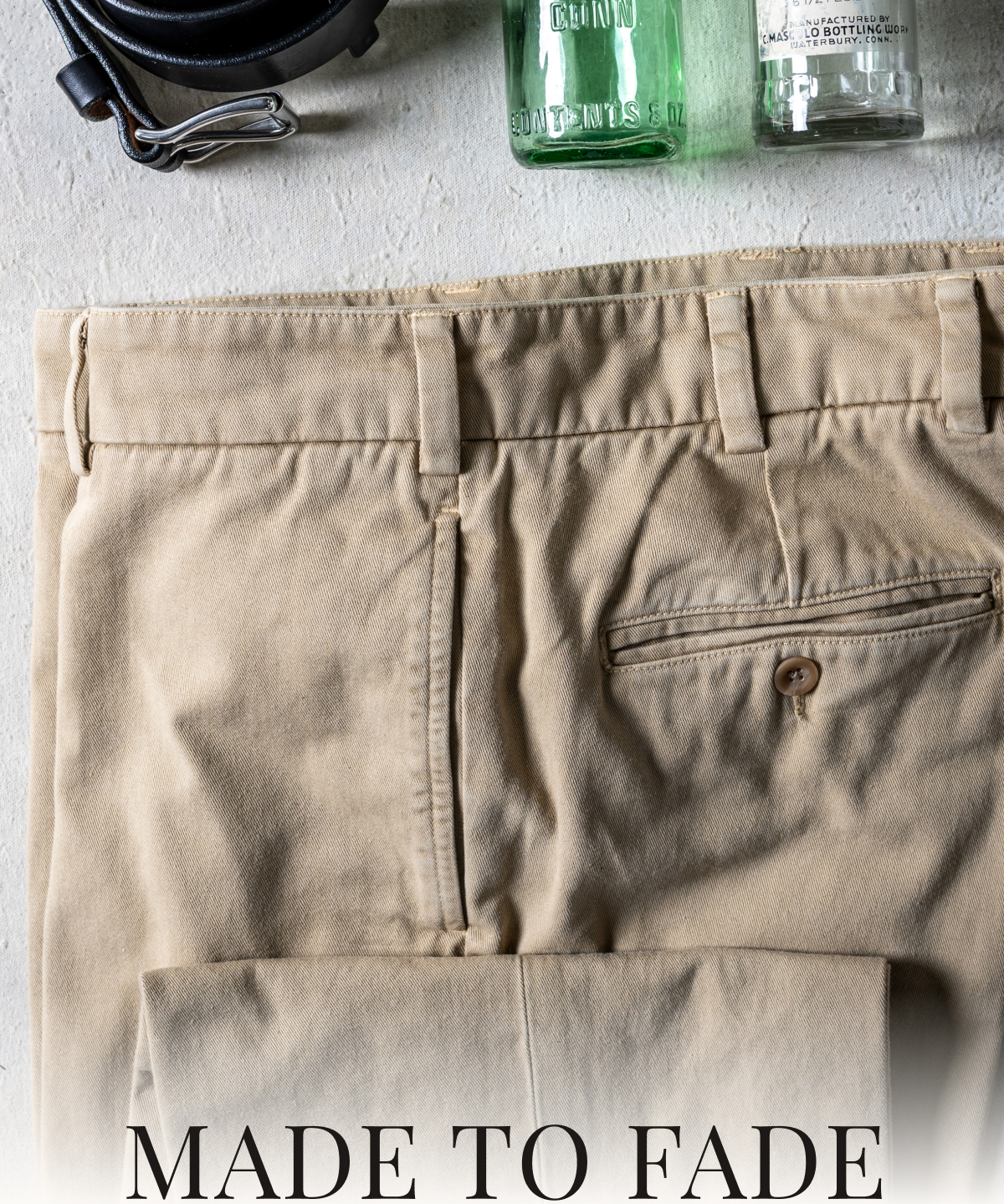 A close up photo of the top portion a pair of khaki Bills Khakis Heritage Wash pants laying on a table. A Bills Khakis belt and some vintage soda bottles frame the pants. Text reads: Made to Fade