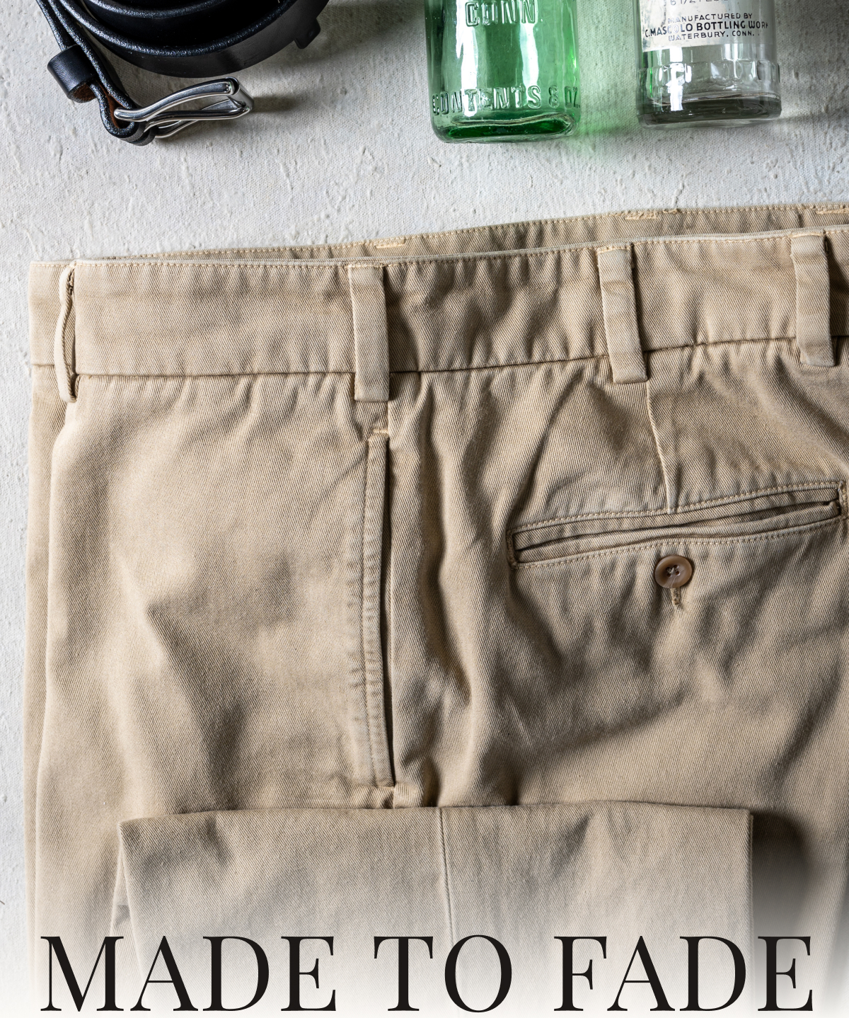 A close up photo of the top portion a pair of khaki Bills Khakis Heritage Wash pants laying on a table. A Bills Khakis belt and some vintage soda bottles frame the pants. Text reads: Made to Fade