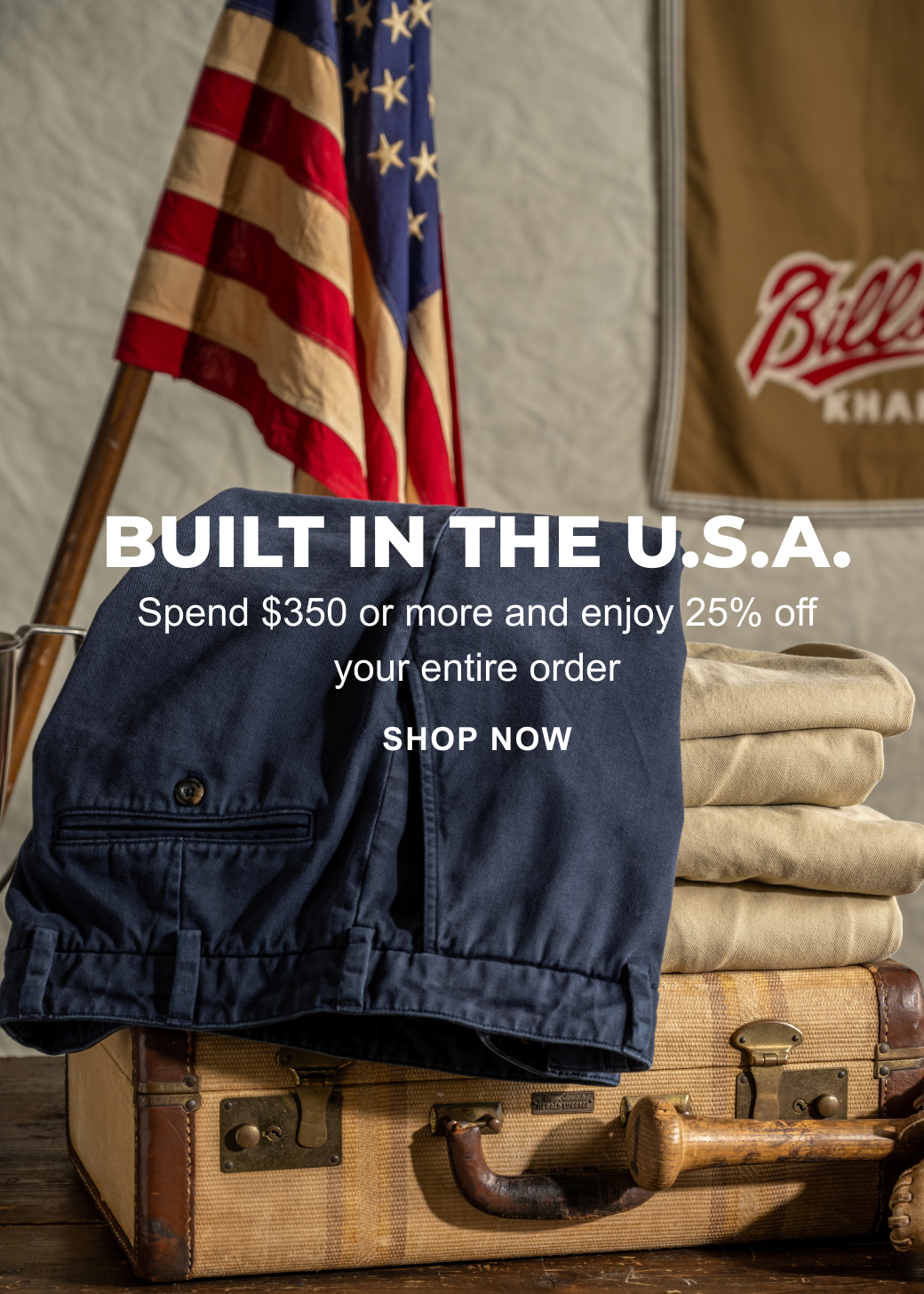 A text overlay reads: Built in the U.S.A. Spend $350 or more and enjoy $25% off your entire order. Shop Now. The image behind the text showcases a pair of navy Bills Khakis khaki pants hanging across a stack of Bills Khaki khaki colored pants on a vintage suitcase on a vintage wooden table. A Bills Khaki flag and an American Flag are displayed proudly in the background.