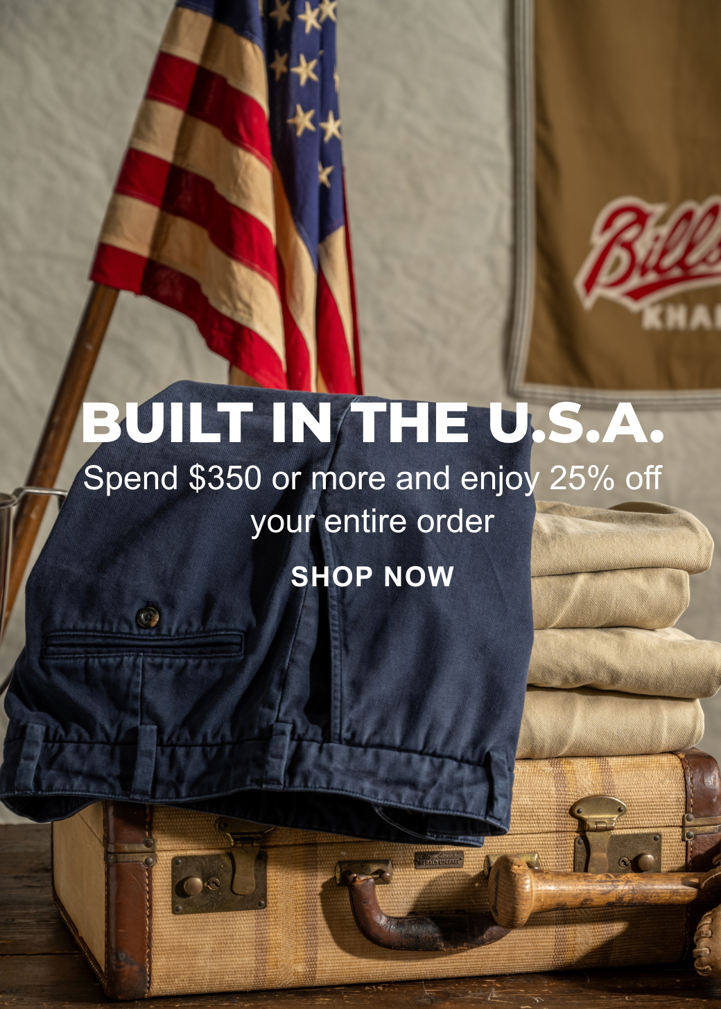 A text overlay reads: Built in the U.S.A. Spend $350 or more and enjoy $25% off your entire order. Shop Now. The image behind the text showcases a pair of navy Bills Khakis khaki pants hanging across a stack of Bills Khaki khaki colored pants on a vintage suitcase on a vintage wooden table. A Bills Khaki flag and an American Flag are displayed proudly in the background.