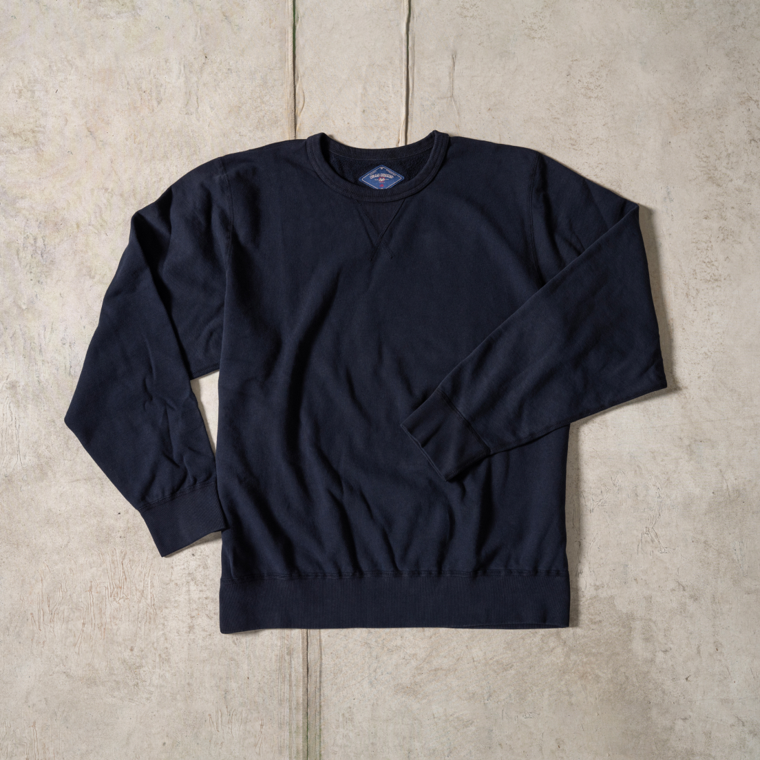 A navy Bills Khaki's Cotton Crewneck laid flat on a tan surface