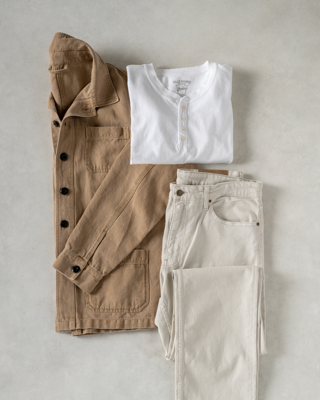 A lay down of a khaki Bills Khaki over shirt, a white Bills Khaki Henley, and a stone colored Bills Khaki Bullard Twill pants on a cream surface