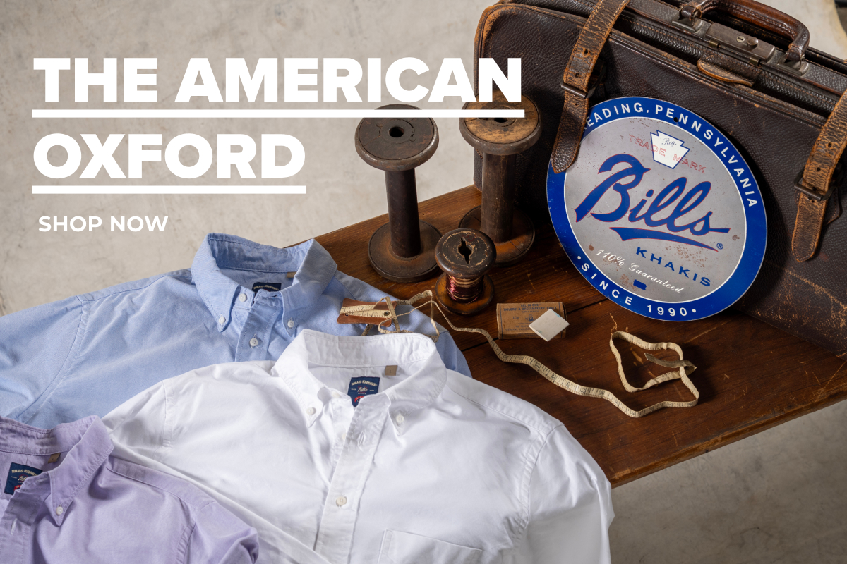 Text Reads: The American Oxford | Shop Now. Text overlays a wooden bench covered with blue, white and purple Bills Khaki American Oxford shirts along with vintage spools, vintage tailors measuring tape, vintage suitcase, and a blue vintage tin signs that reads Bills Khakis
