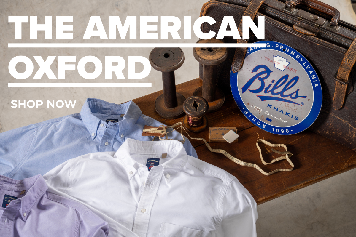 Text Reads: The American Oxford | Shop Now. Text overlays a wooden bench covered with blue, white and purple Bills Khaki American Oxford shirts along with vintage spools, vintage tailors measuring tape, vintage suitcase, and a blue vintage tin signs that reads Bills Khakis