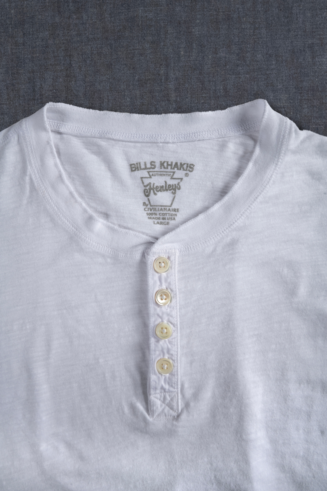 A detail of the color and button area of a white Bills Khakis Henley laying on a grey surface