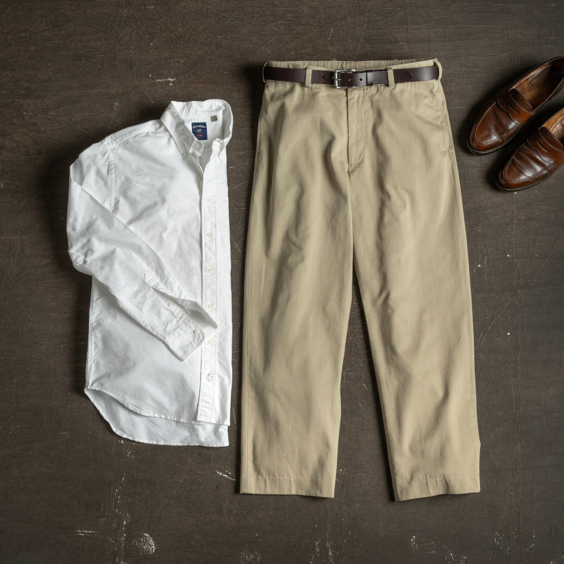 A laydown of Bills Khakis M1 khakis + white standard fit shirting on a dark brown, industrial surface