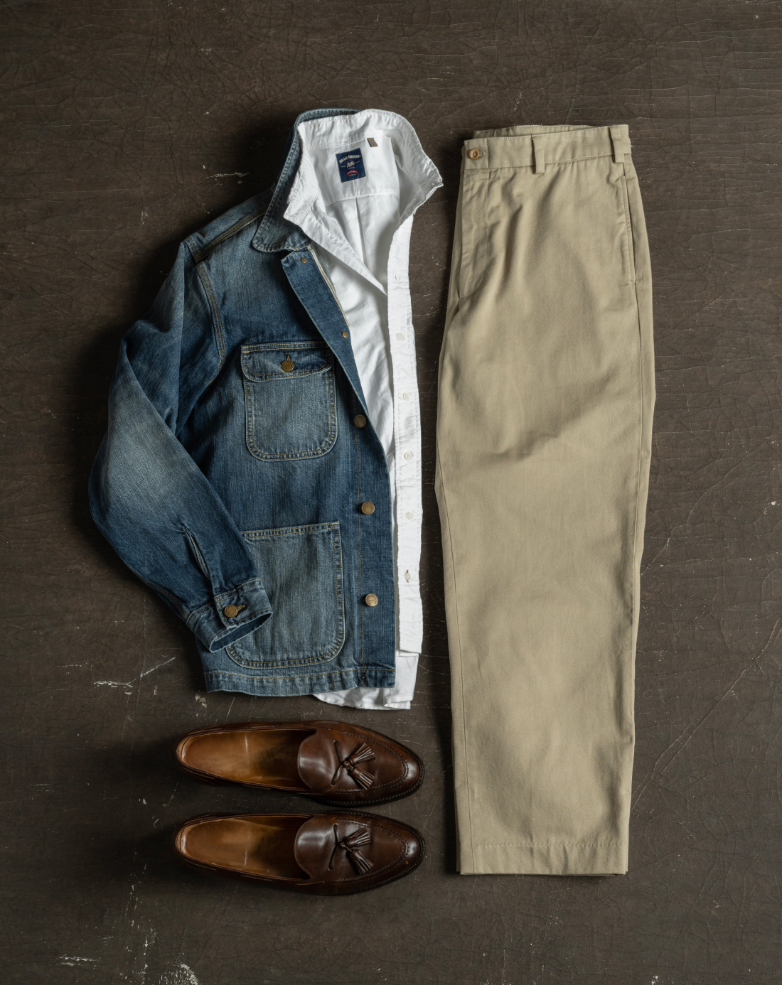An outfit idea laying on a dark brown, industrial surface. The outfit consist of Bills khakis M1 Khakis, a white Standard Fit Shirt, and denim jacket