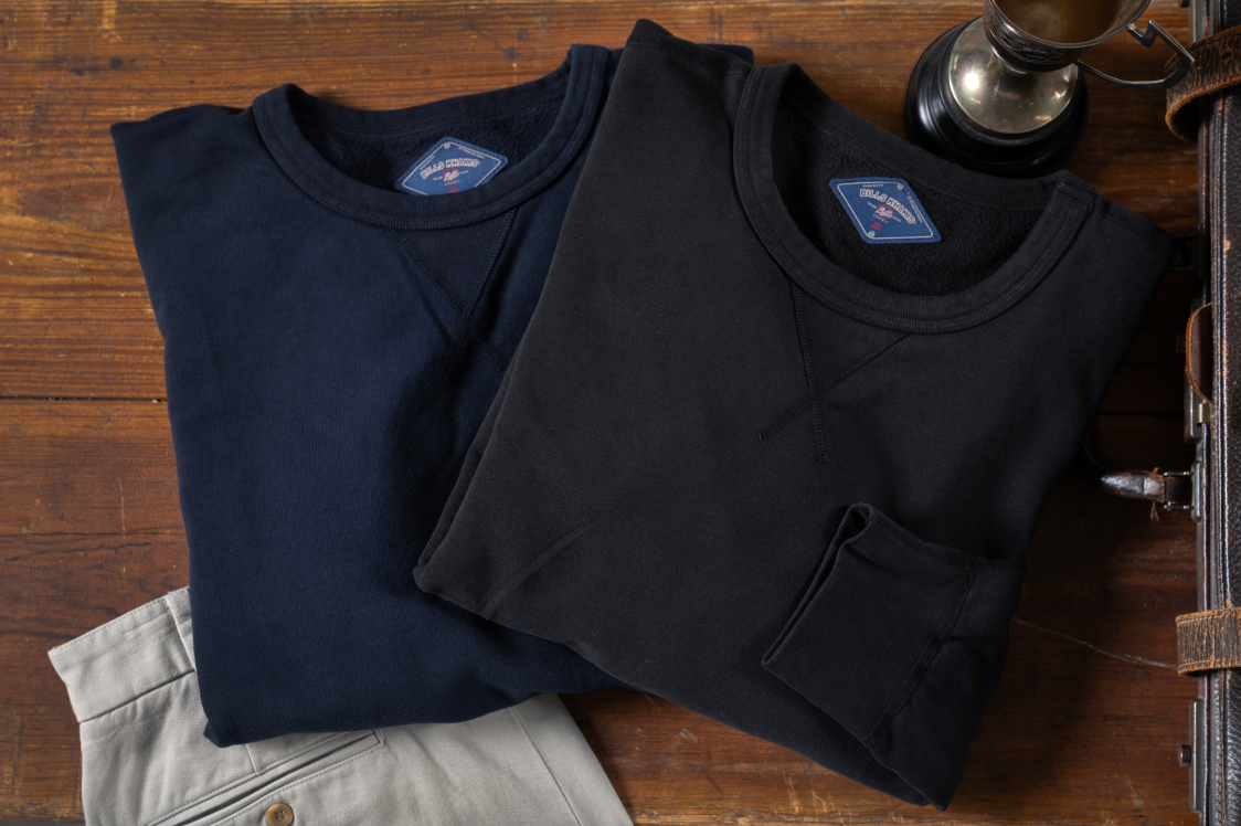 Two Bills Khakis lightweight sweaters sitting folded on a dakr brown wood surface..one in navy and one in black