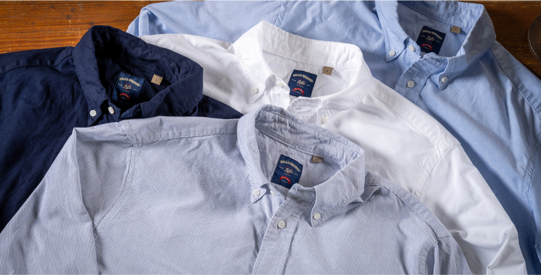 Four Bills Khakis Standard Fit shirts in a spectrum from white to dark navy lay flat along a wooden surface