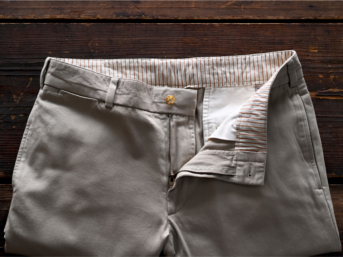 A detail of a stone pair of Bills Khakis Khaki waste and zipper laid on a dark brown surface