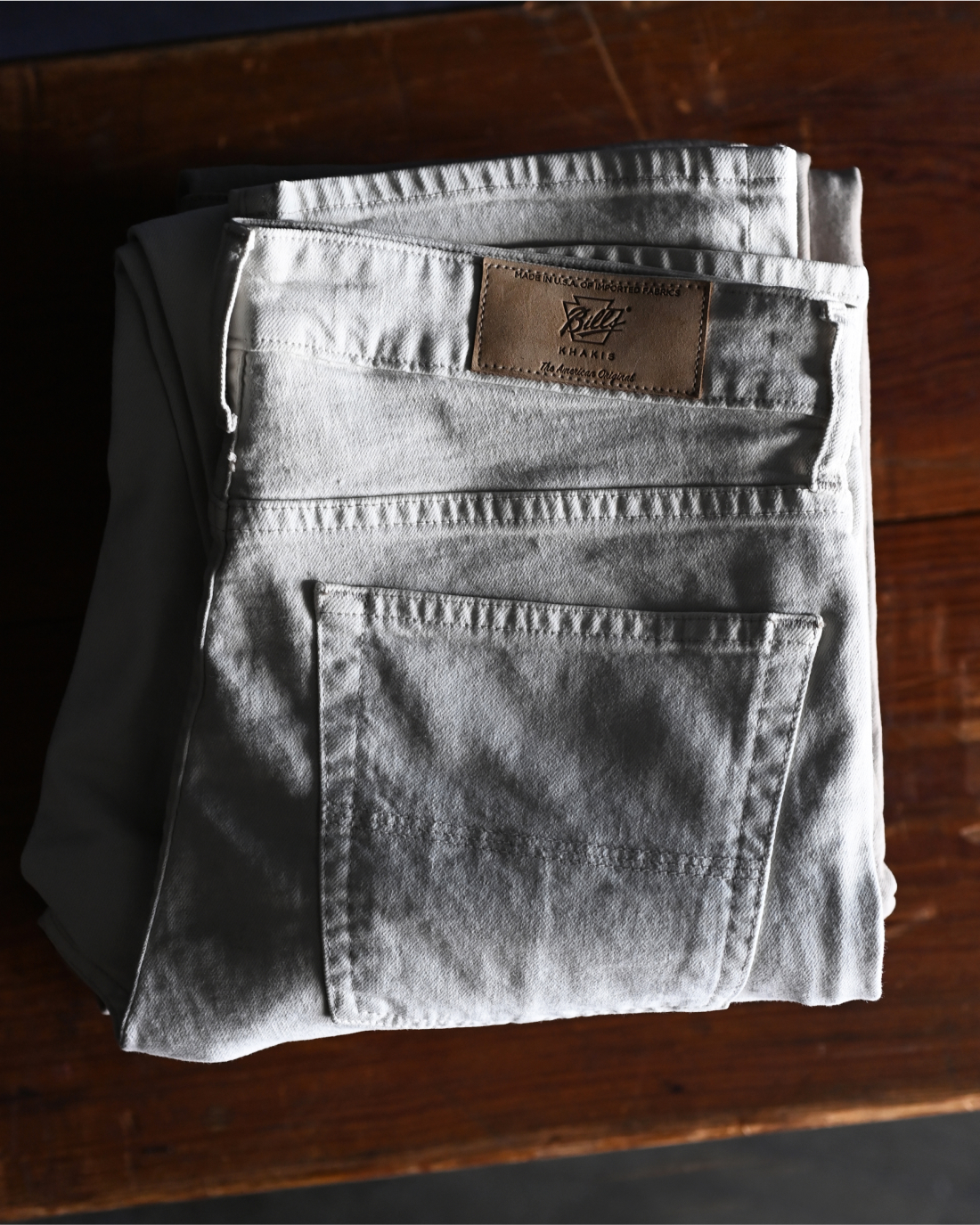 A stone colored pair of Bills Khaki's 5-pocket pants folded on a wooden surface showing the back pocket and tag