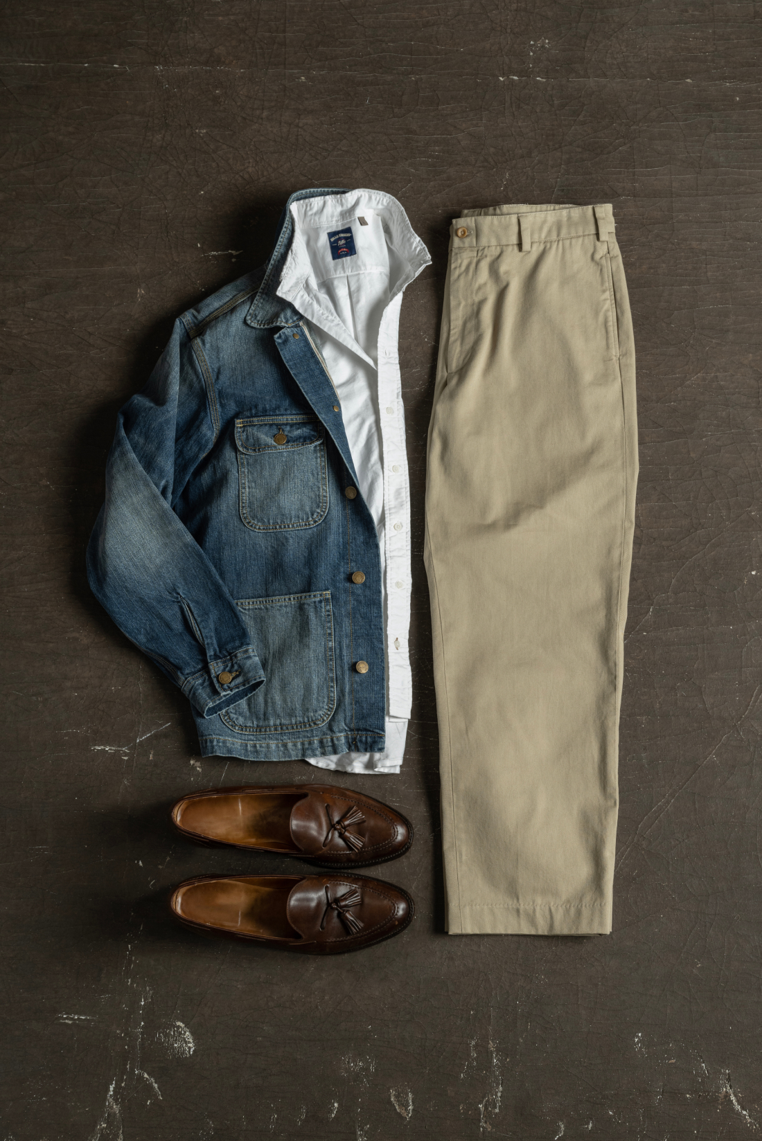 A Bills khaki denim jacket, white standard fit shirt, and khaki pants sit with a pair of leathe rloafers on an industrial brown surface