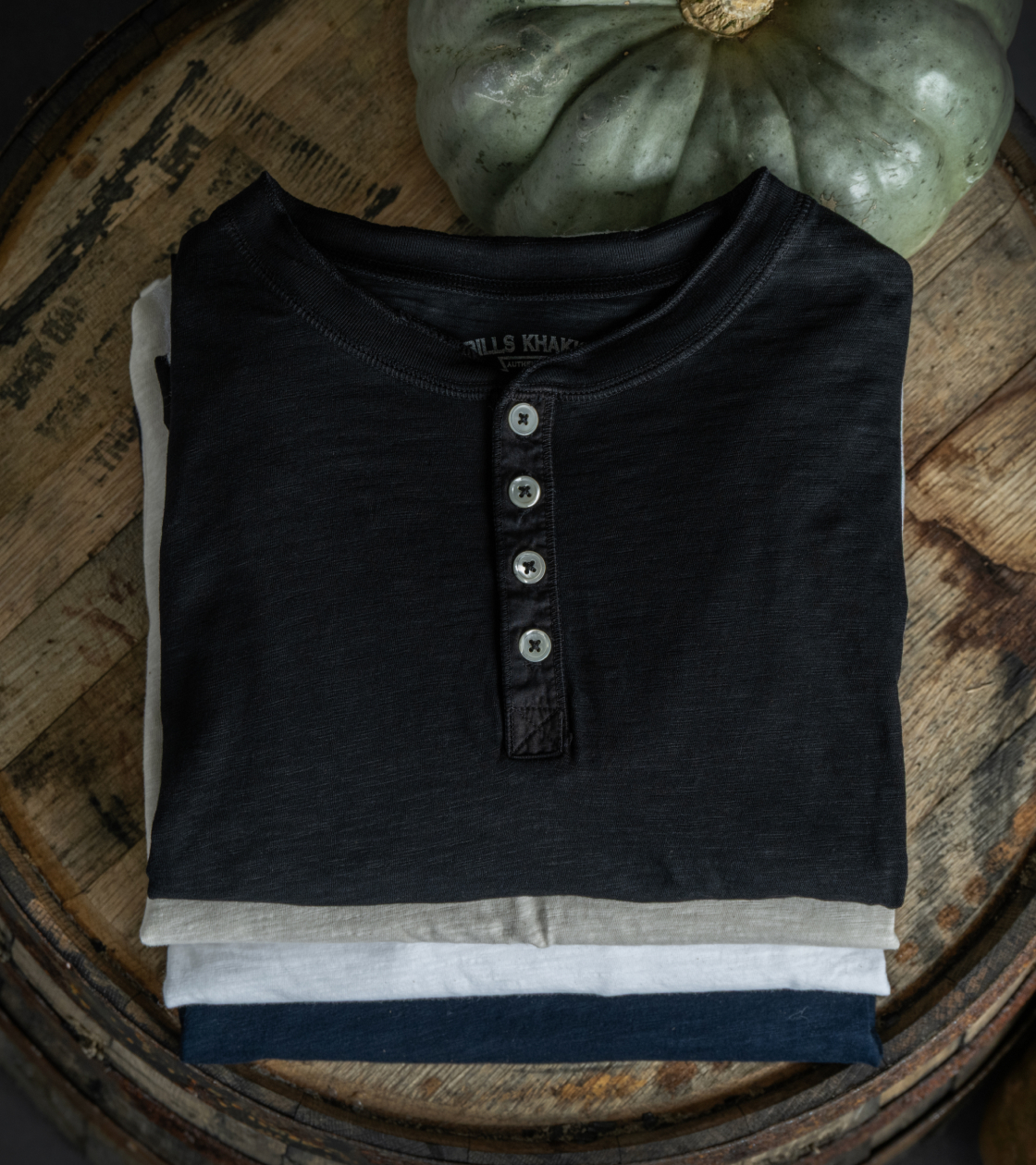 Bills Khaki Henleys sit folded and stacked on a vintage wood barrel alongside a green pumpkin