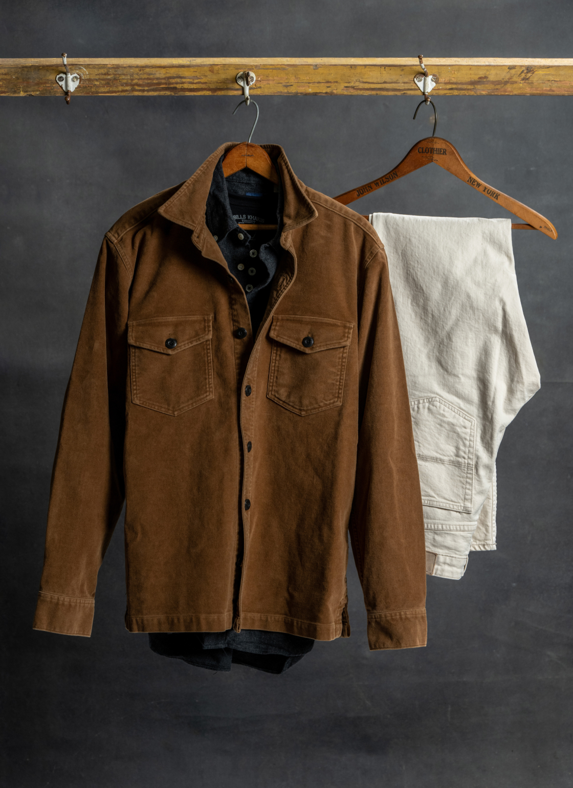 Against a dark backdrop a Bills Khaki Weathered Moleskin Chore Jacket hangs slongside a dark Standard Fit shirt and stone 5 pocket pants