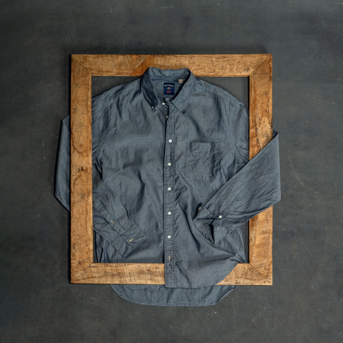A grey Bills Khakis Standard Fit Shirt lays on an industrial black surface along with some wooden accents