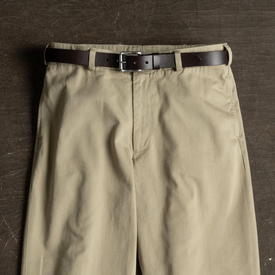 A pair of khaki Bills Khakis along with a Bills Khakis belt lay on an industrial brown surface zipper up