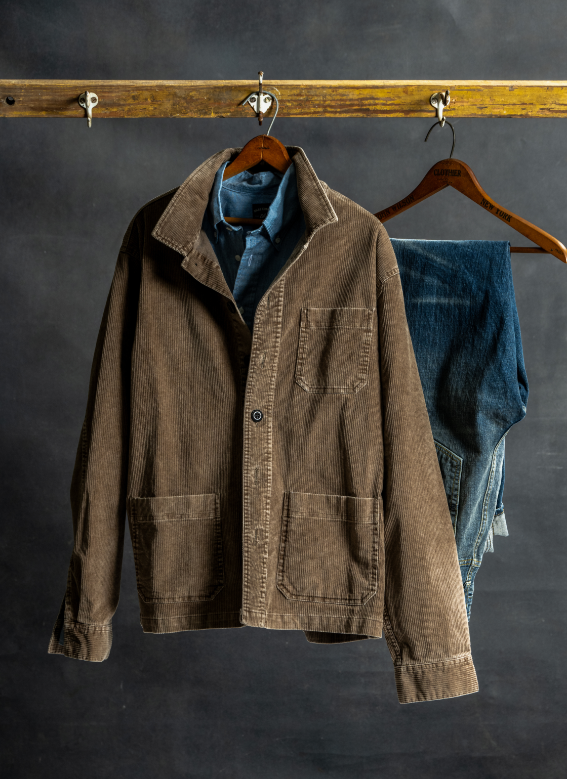 A Bills Khakis brown cordoroy Chore jacket hangs over a blue standard fit shirt and alongside Bills denim pants against a black industrial backdrop