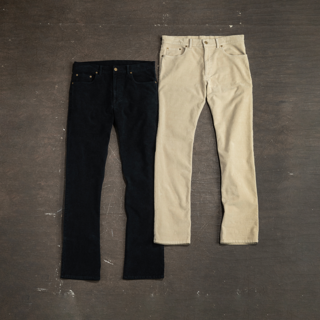 A black pair and a light khaki pair of Bills Khaki moleskin pants lay alongside each other on a brown industrial surface