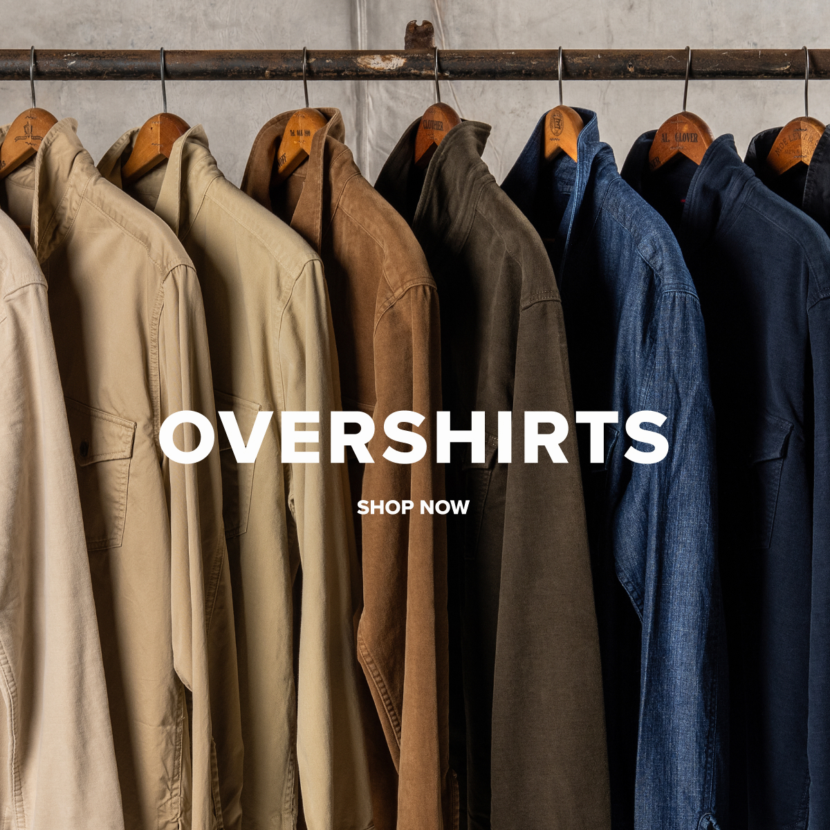 An array of Bills khaki overshirts from light khaki to brown to dark navy and black hang from an industrial pole and hangers. Text reads: Overshirts | Shop Now