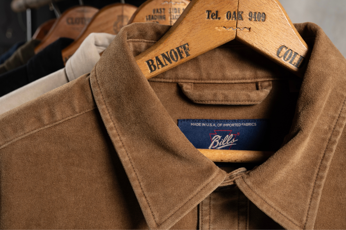 A close up of a brown Bills Khaki chore jacket collar and Bills Khaki tag hanging from a hangar showing off its stitching