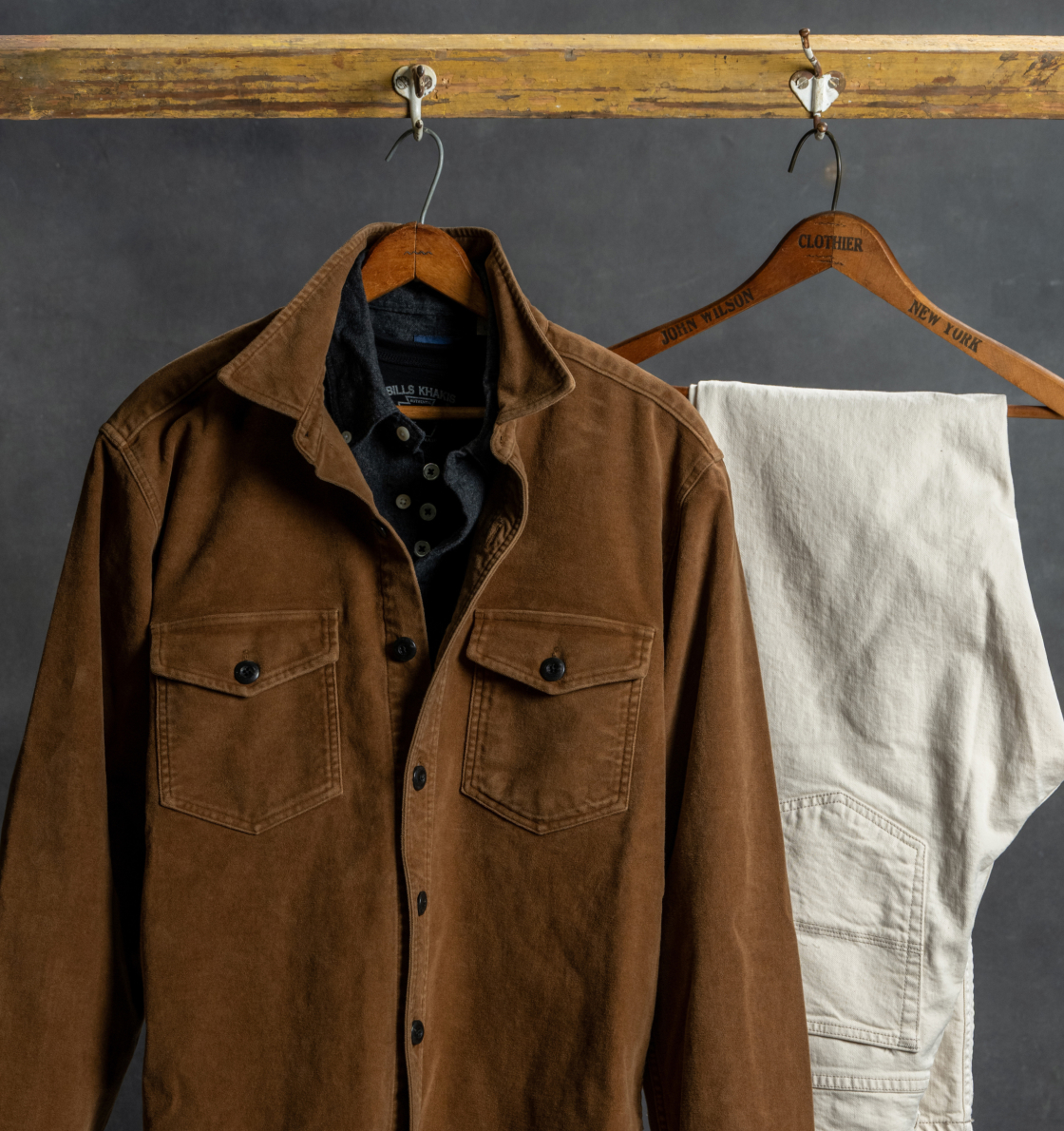 A brown Bils Khaki chore jacket over a navy Bills Khaki standard fit shirt alongside hanging next to a pair of 5 pocket Bills Khaki pants against a dark industrial background