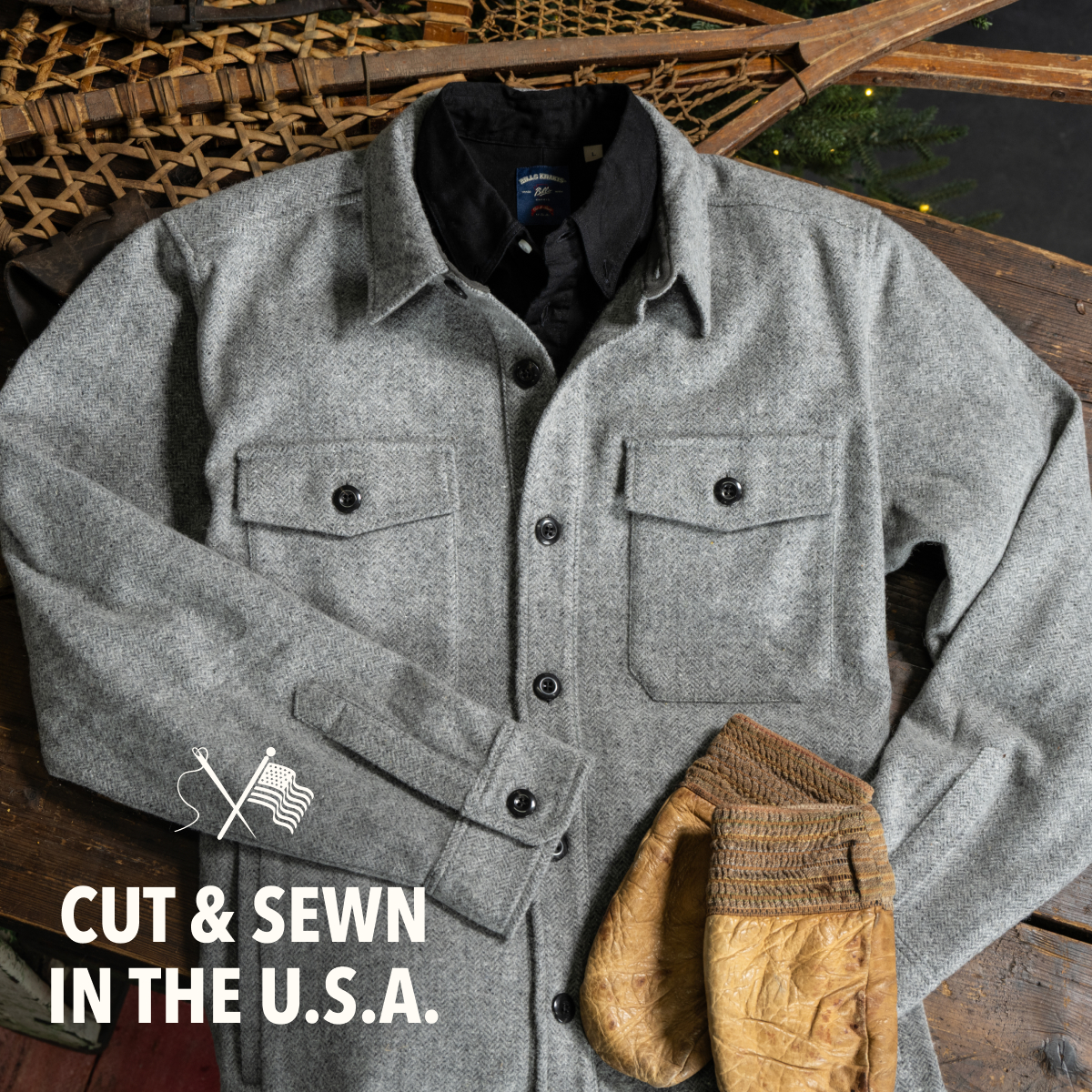 A new Bills Khakis Wool Shirt sits outside a Bills Khaki standard fit shirt on a worn, dark wooded surface. Off the crop are vintage gloves, snow shoes and eveidence of some pine branches
