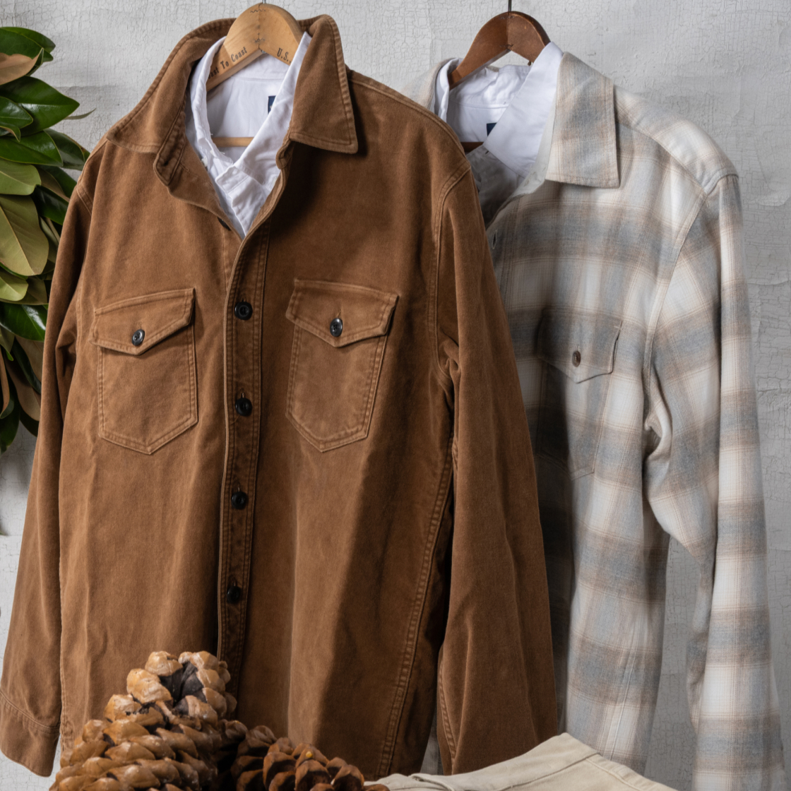 A new Bills Khaki Ombre Flannel hangs alongside a Bills Khaki Moleksin Overshirt. In the surroundings are evidence of pinecones and a holiday wreath