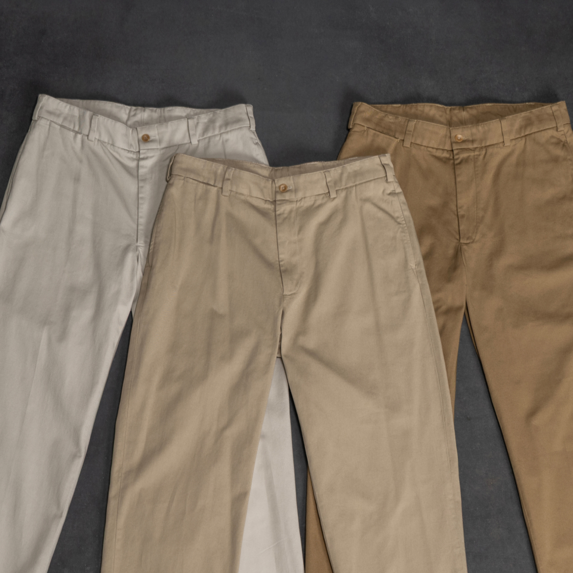 Three pairs of Bills Khakis legendary Original Twill Khakis in tones from stone to khaki lay on an grey industrial surface