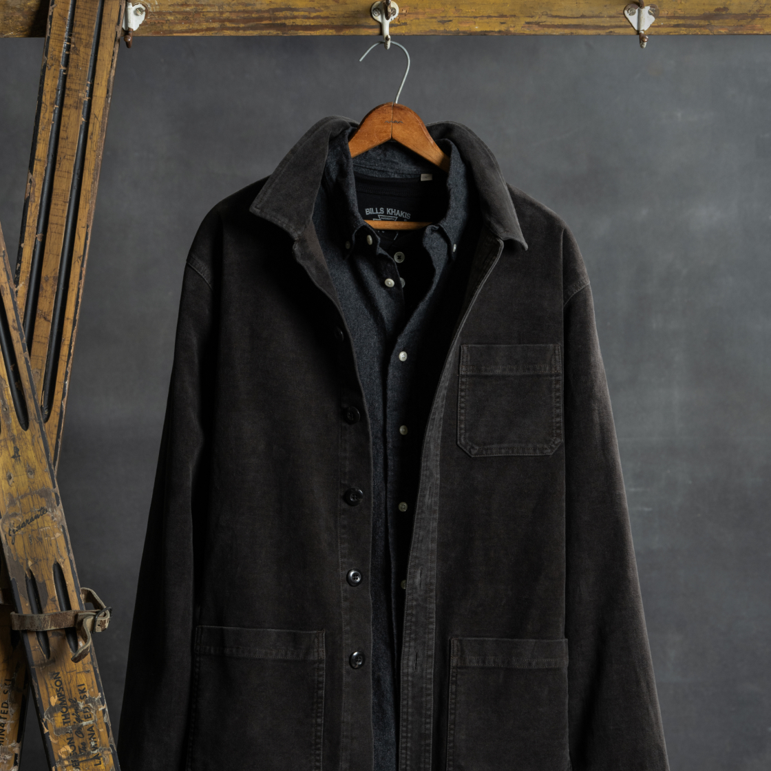A dark Bills Khaki jacket sits over an Bills Khaki shirt while hanging from a hangar alongside vintage skis against a dark backdrop