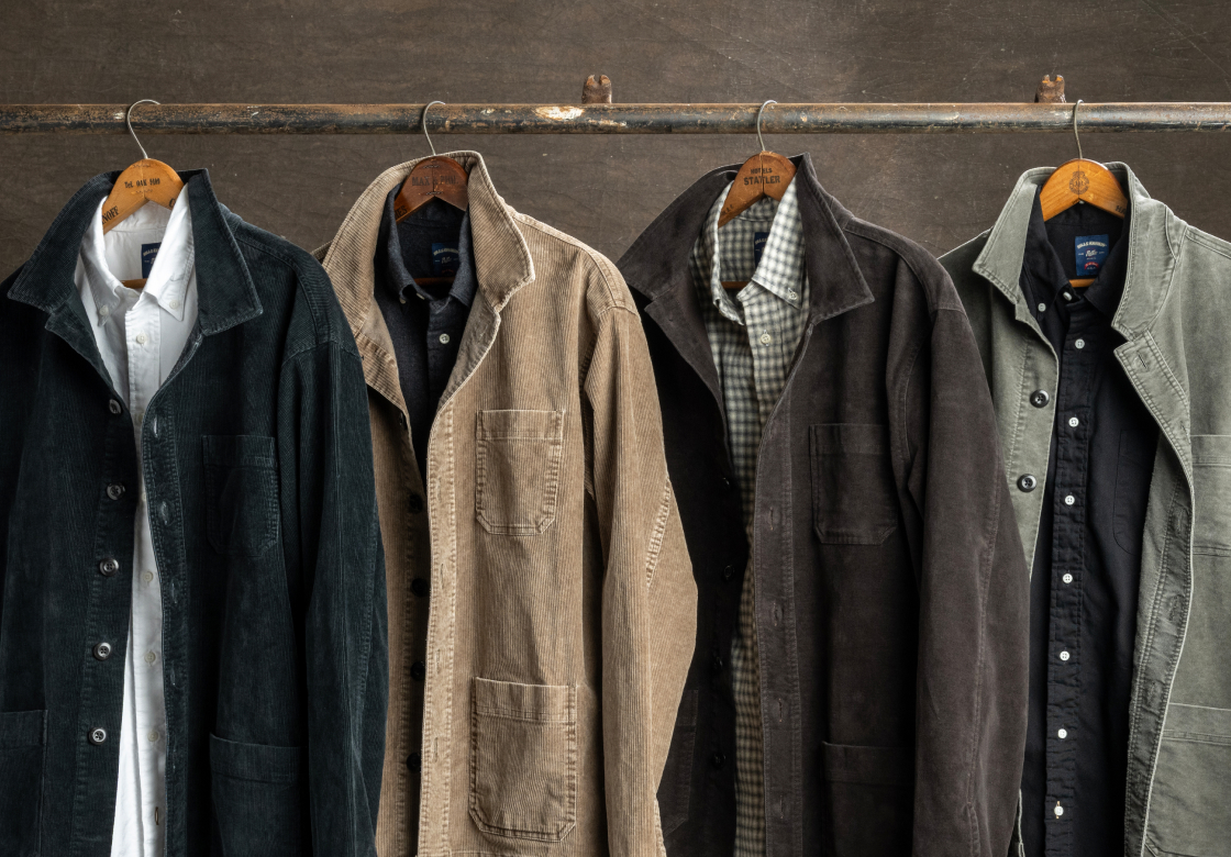 Four Bills Khakis overcoats hang over Bills Khaki standard fit shirts from hanagers off a metal rod against a brown industrial surface.