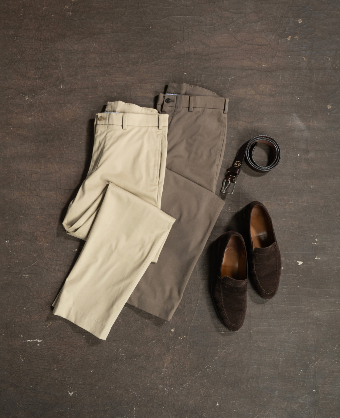 Bills Khakis travel twill pants in two khaki tones lay against an industrial surface alognside a Bills Khakis belt