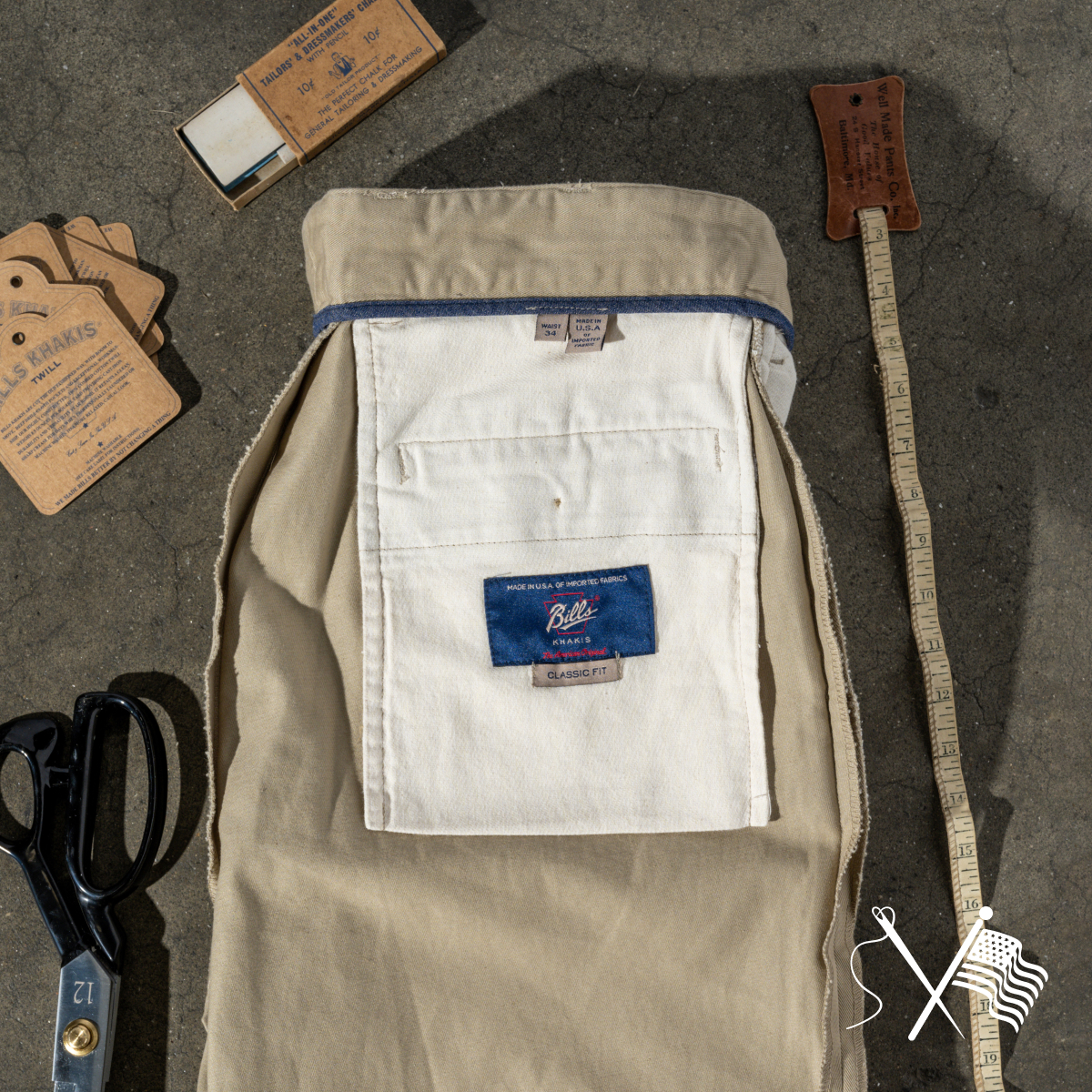 A pair of Bills Khakis khaki pants lay inside-out displaying the authentic stitch work on an industrial stone surface. Around the pants are a vintage measuring tape, scissors, tags and thread and needle