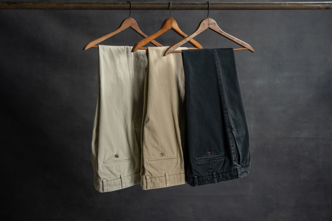 Three pairs of Bills Khakis khaki pants hang from hangars on an industiral pole against a dark backdrop