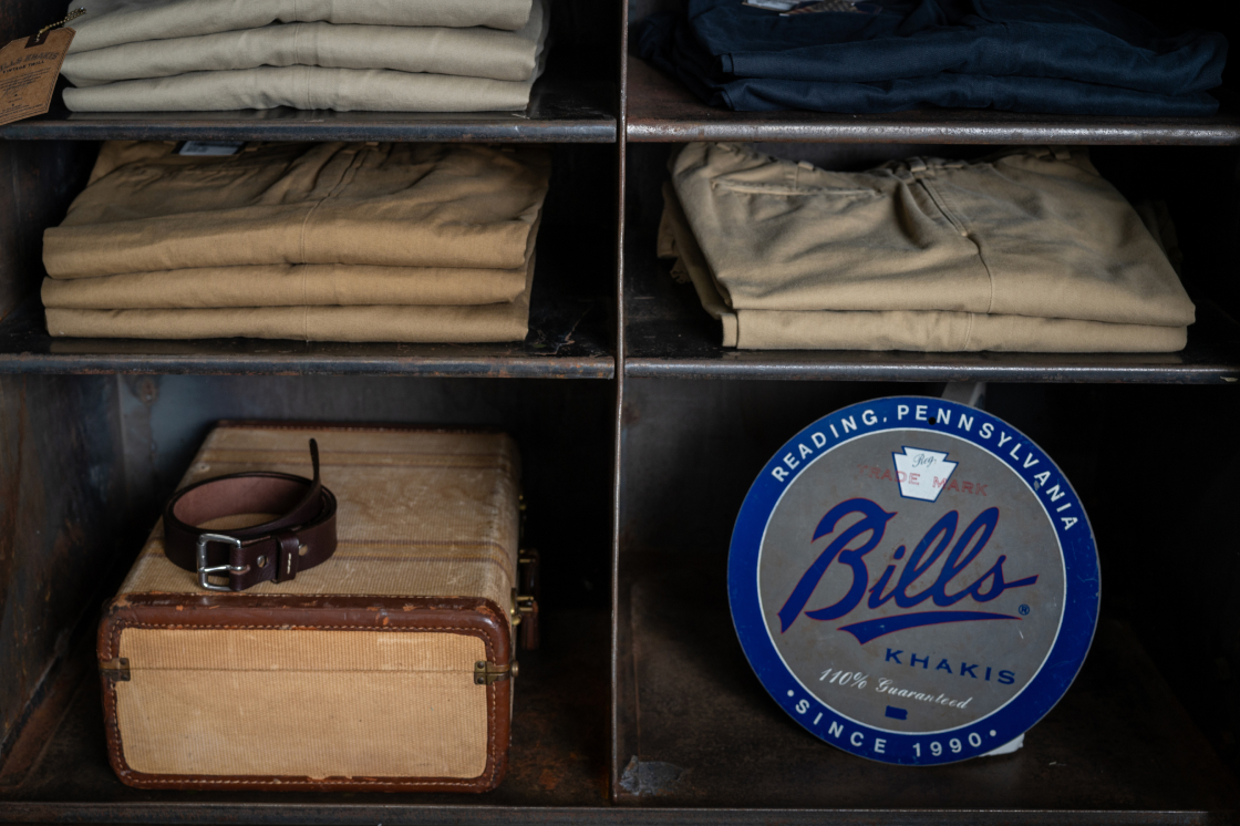 An aged, industrial shelving unit displays many pairs of folded Bills Khakis pants as well as vintage suitcase and a vintage Bills Khakis tin sign