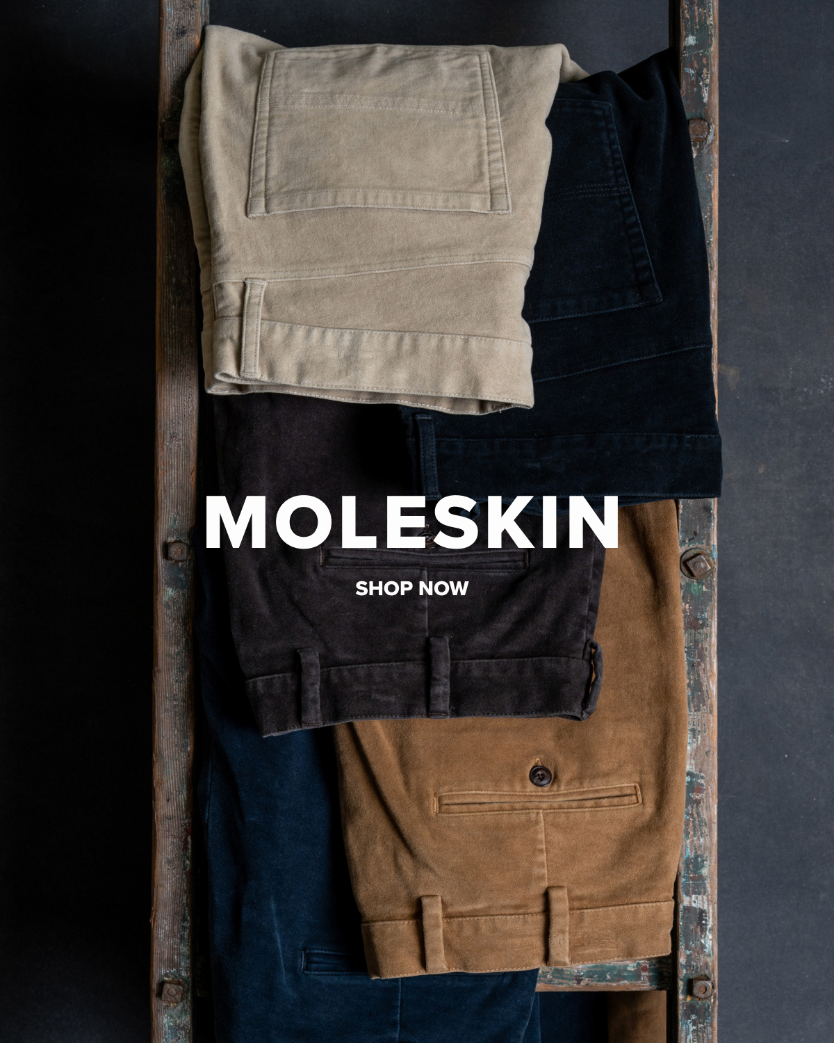 Five pairs of Bills Khakis Moleskin pants hang from an aged, vintage ladder against a dark backdrop.