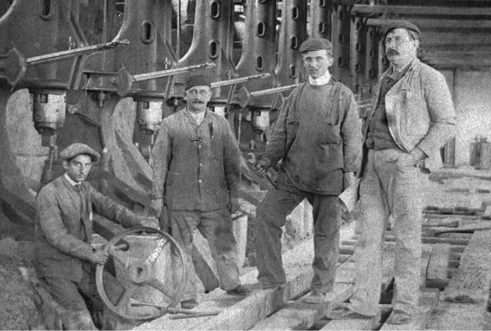 Four men stand in an industrial warehouse job site in the late 1800s/early 1900s wearing chore jackets