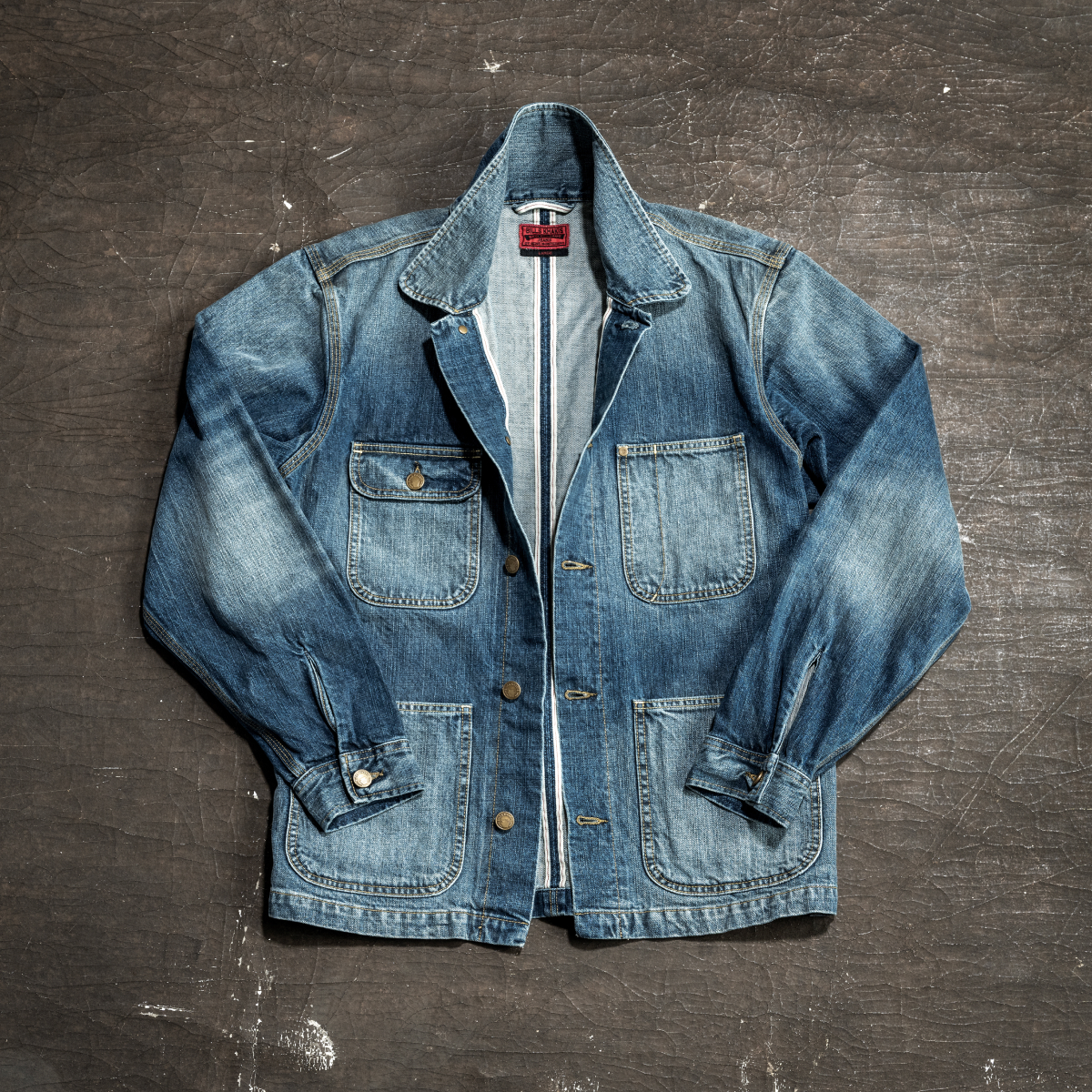A Bills Khakis denim chore jacket lays against an inustrial surface.