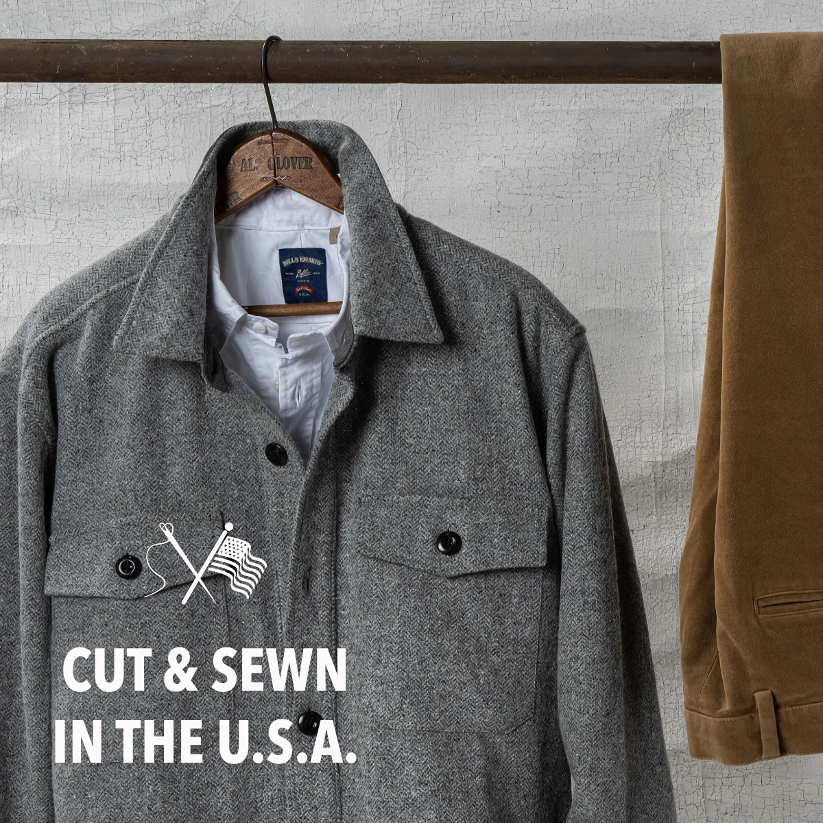 A Bills Khaki Wool Herringbone jacket hangs from an aged, rusted metal pole. A pair of Bills Khakis moleskin pants hang alongside it