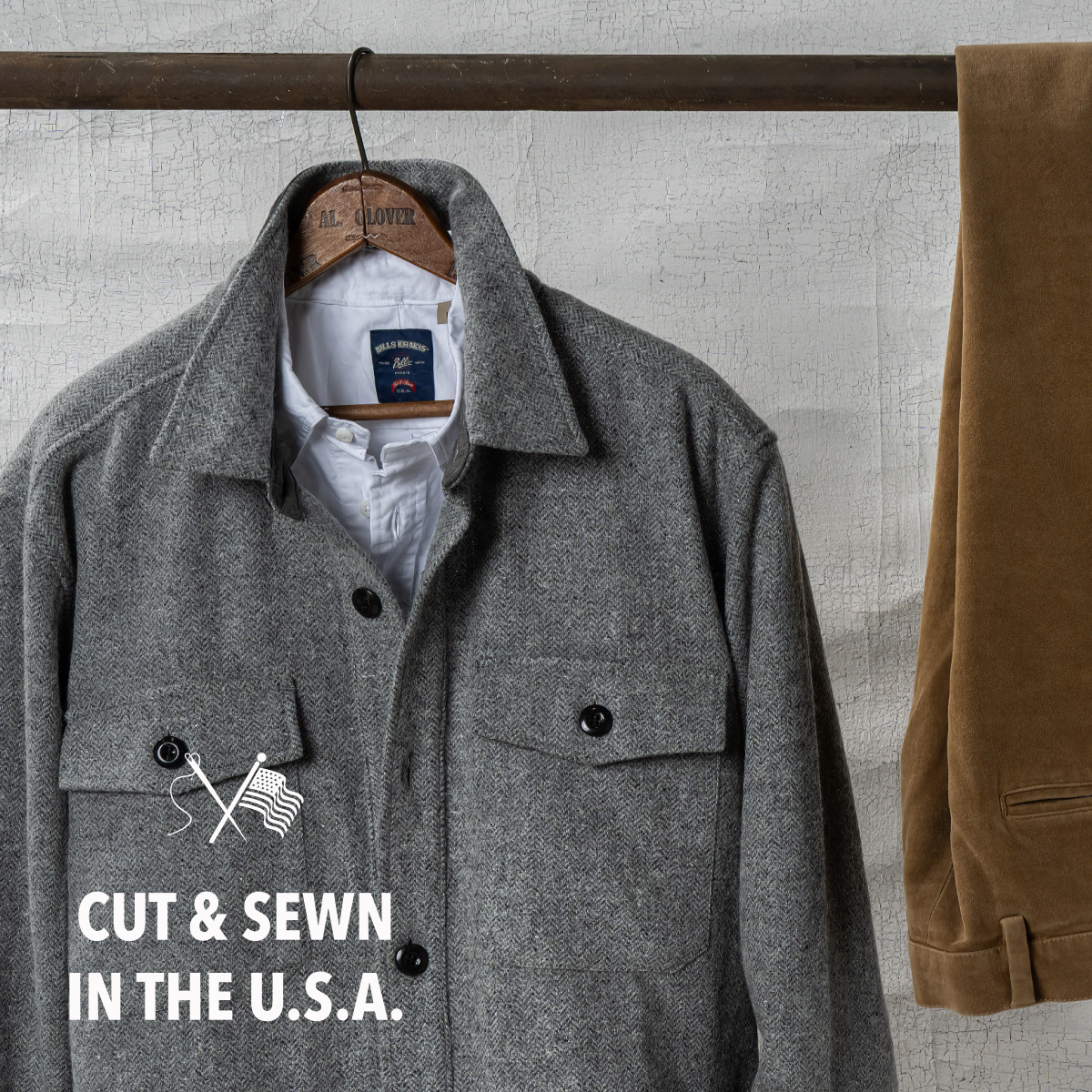 A Bills Khaki Wool Herringbone jacket hangs from an aged, rusted metal pole. A pair of Bills Khakis moleskin pants hang alongside it