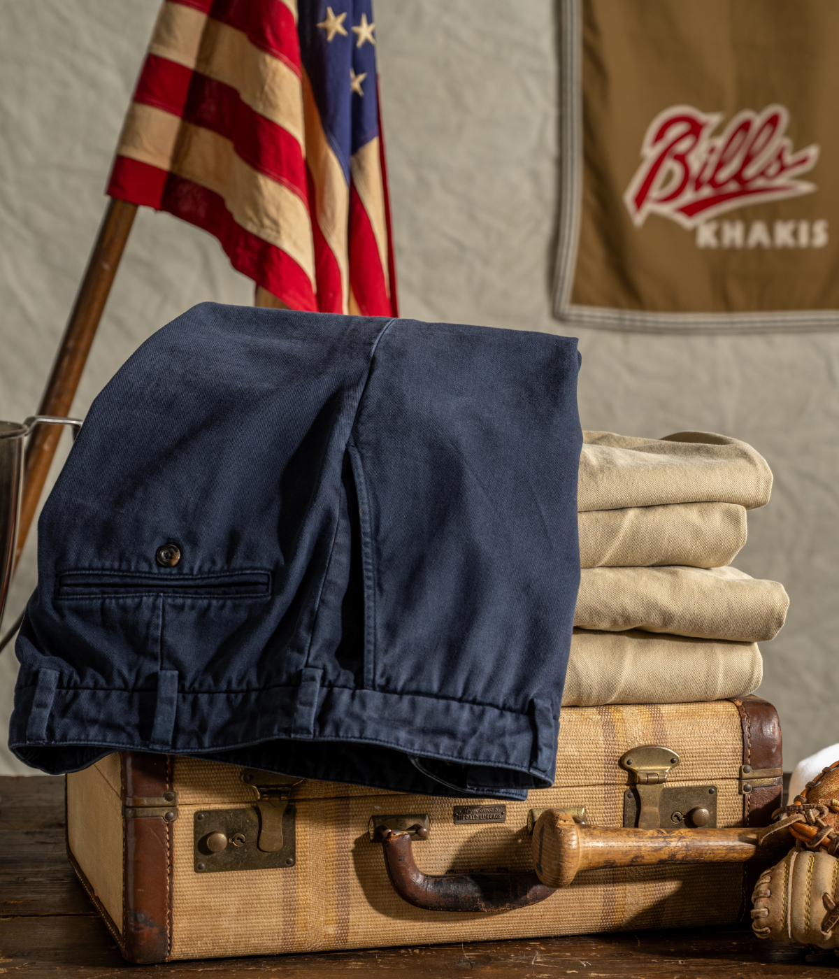 A photo of Bills Khakis Heritage Wash pants in khaki and navi folded on a vintage suitcase with an American flag and vintage Bills decal in the background