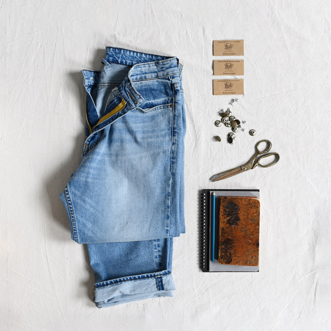 A pair of Bills Khakis denim lays on a white cloth surface. Scissors, Sketchbooks, loose pants rivets, and unsewn Bills Khaki back waist tags.