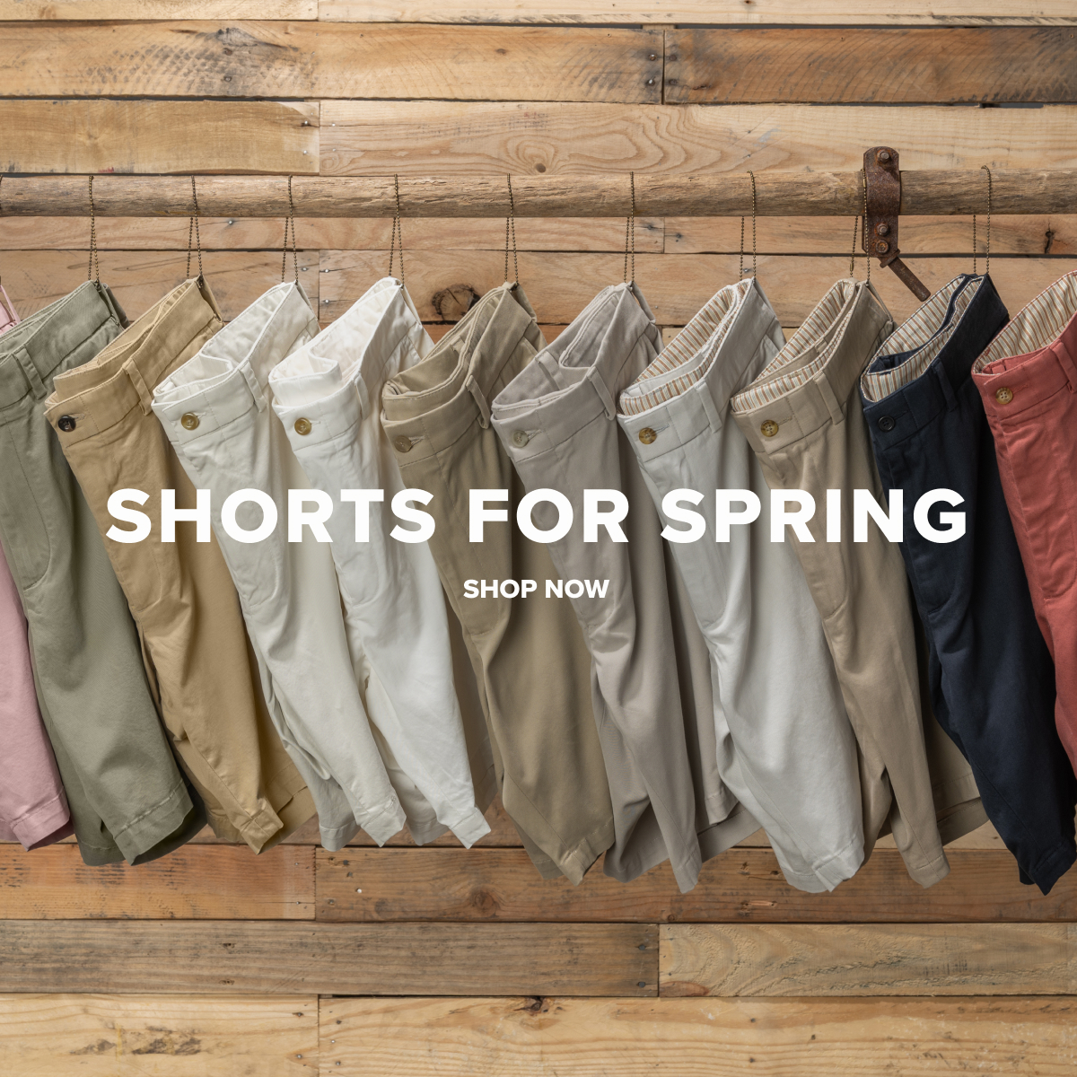 An array of Bills Khakis Shorts hanging from a wooden pole, against a wooden backdrop. Text reads: Shorts for Spring | Shop Now