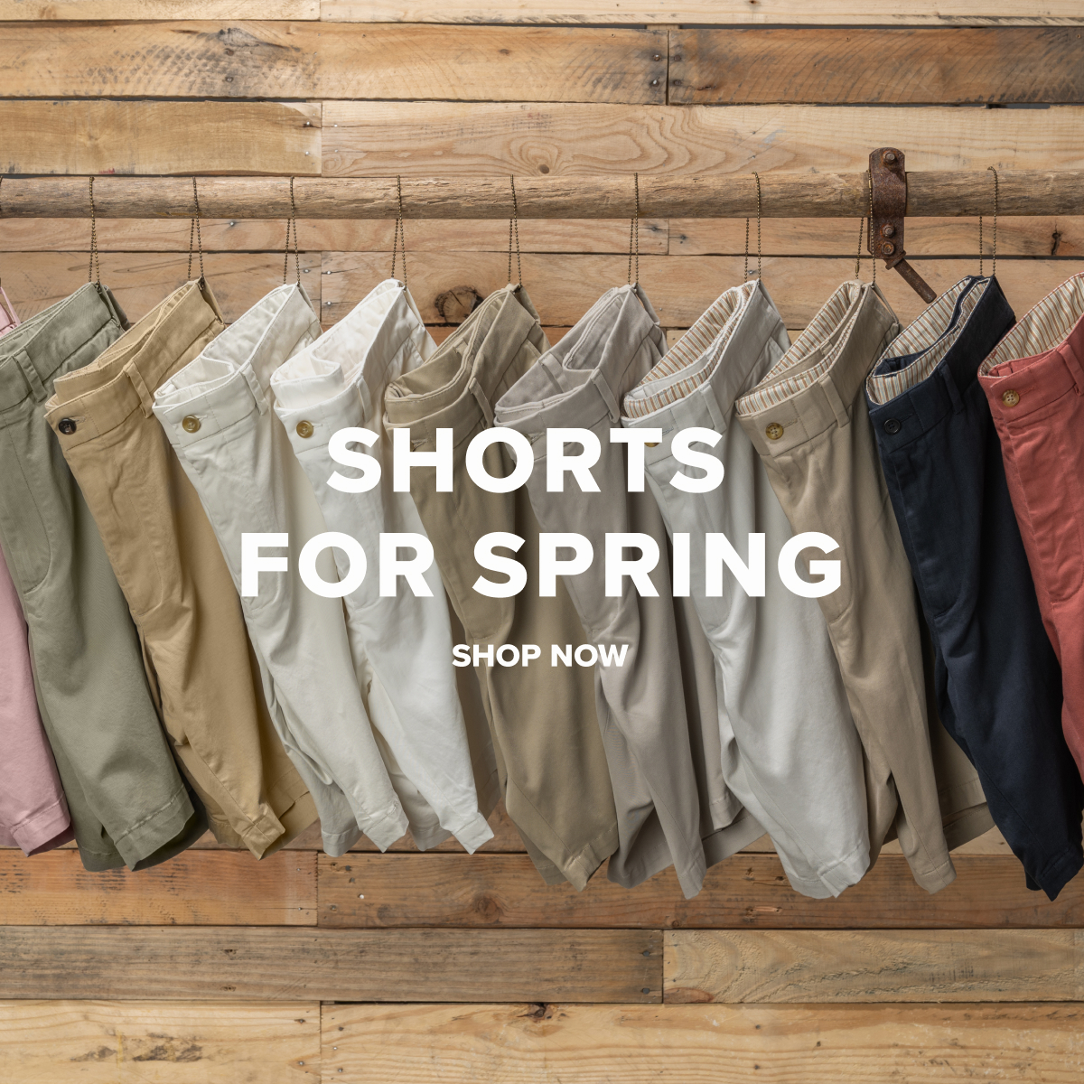 An array of Bills Khakis Shorts hanging from a wooden pole, against a wooden backdrop. Text reads: Shorts for Spring | Shop Now