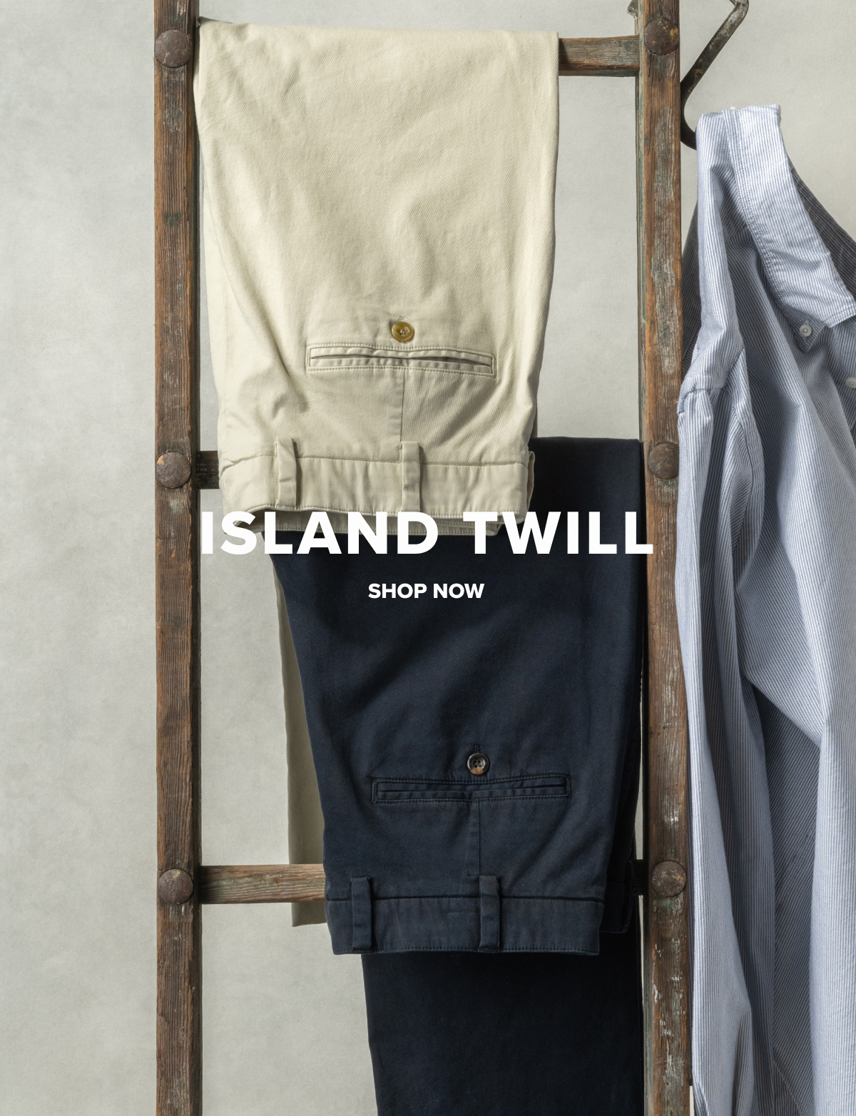 Two pairs of Island Twill – Classic Fit – M2 pants in Khakis and Midnight hang from a vintage ladder along with a Bills Khakis Standard Fit Shirt. Text overlay reads: Island Twill | Shop Now