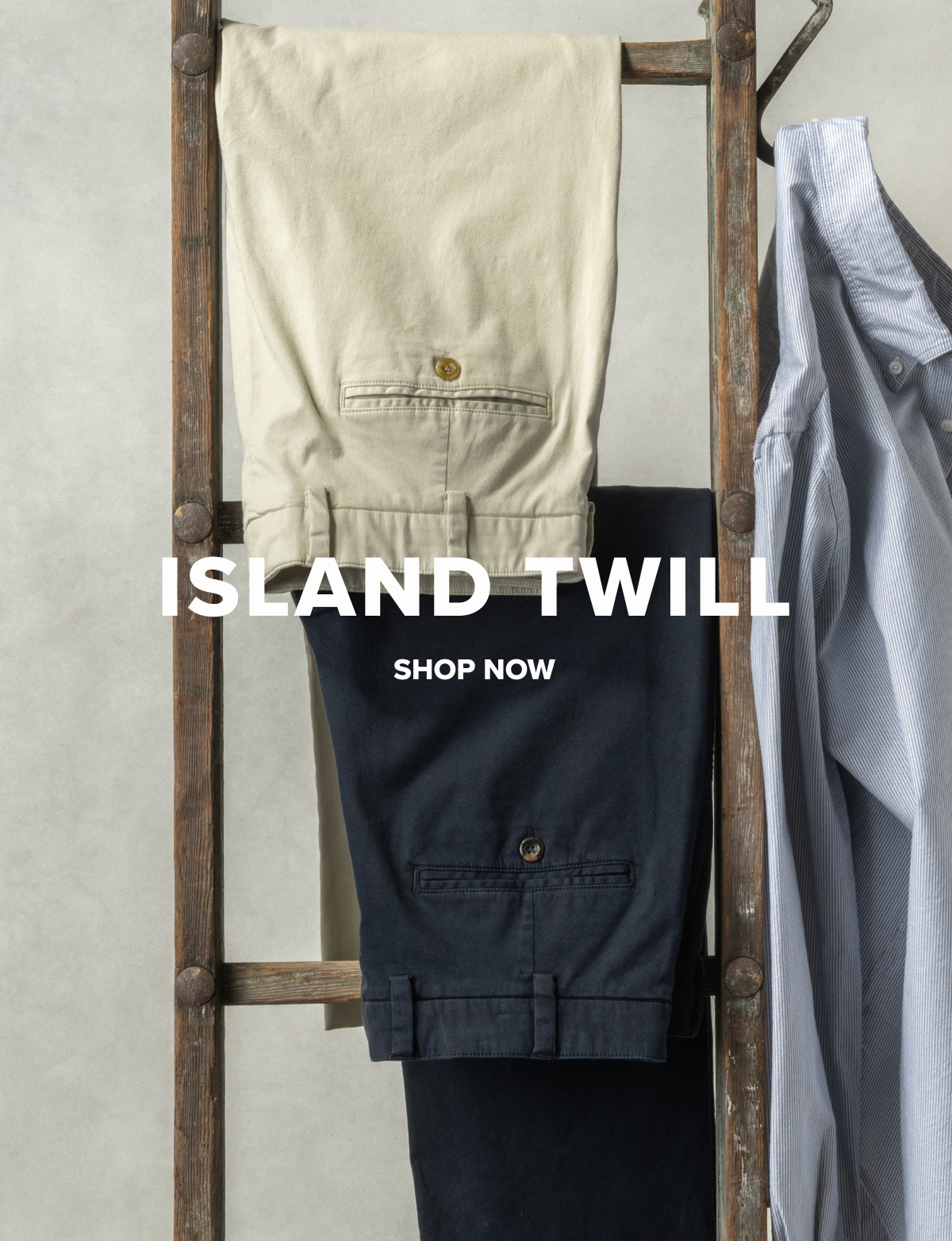 Two pairs of Island Twill – Classic Fit – M2 pants in Khakis and Midnight hang from a vintage ladder along with a Bills Khakis Standard Fit Shirt. Text overlay reads: Island Twill | Shop Now