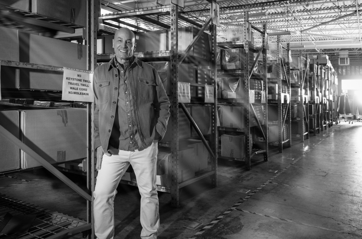 An image of Bills Khakis president Ed Mascolo smiling in his Bills Khakis daily wear standing amongst shelves of product in the Bills Khakis warehouse
