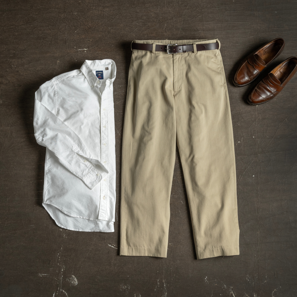 A pair of Bills Khakis Original Twill pants and a white Stand Fit shirt lay alongside leather loafers on a dark, warm industrial colored surface